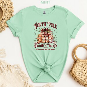 A mint green t-shirt with a Christmas-themed graphic design, including a stack of presents, a gingerbread man, and other festive elements. The t-shirt is displayed on a white background, along with a woven basket and a straw hat.