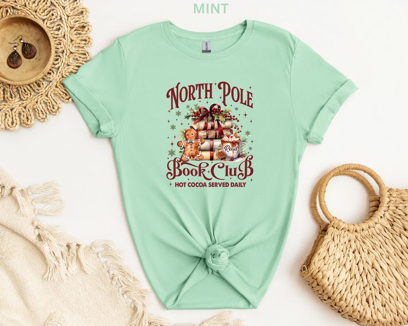 A mint green t-shirt with a Christmas-themed graphic design, including a stack of presents, a gingerbread man, and other festive elements. The t-shirt is displayed on a white background, along with a woven basket and a straw hat.