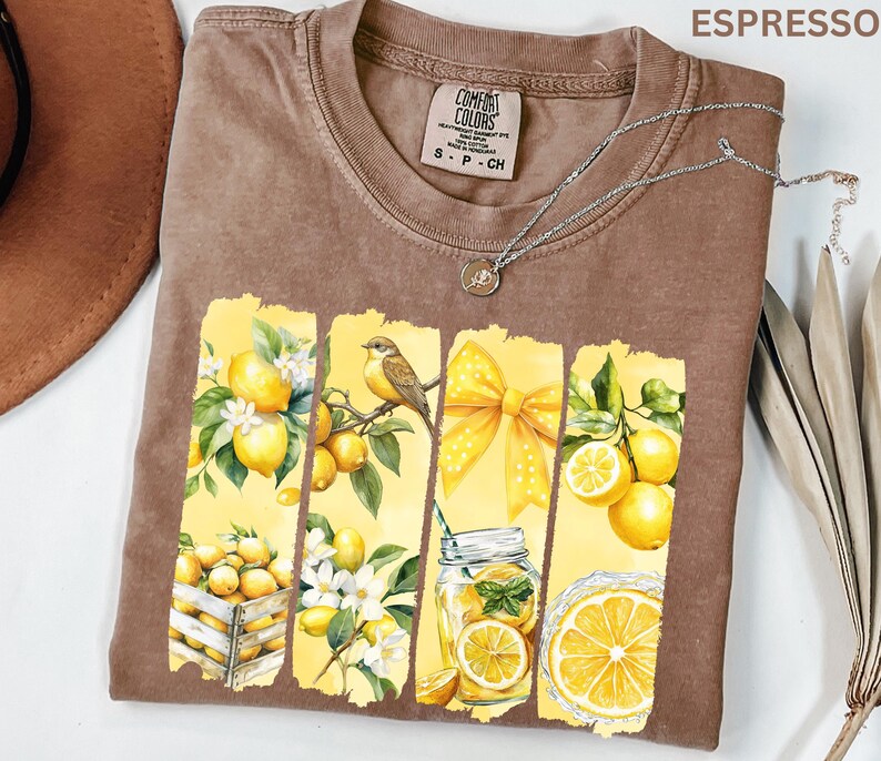 May include: Espresso-colored Comfort Colors t-shirt featuring a lemon-themed graphic. The design includes lemons, a bird, a yellow bow, and a jar of lemonade. A silver necklace with a pendant is draped across the shirt.