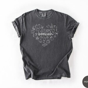May include: A charcoal gray short-sleeve t-shirt featuring a white graphic design in the shape of a heart. The design includes the word bookish in the center surrounded by small line art icons of books, pumpkins, mugs, leaves, and winter accessories.