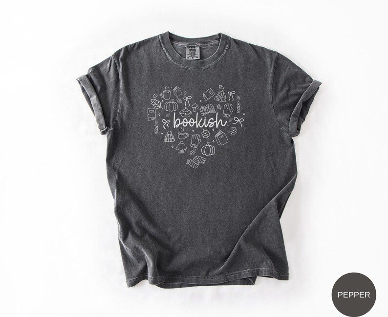 May include: A charcoal gray short-sleeve t-shirt featuring a white graphic design in the shape of a heart. The design includes the word bookish in the center surrounded by small line art icons of books, pumpkins, mugs, leaves, and winter accessories.