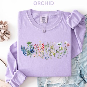 May include: A light purple long-sleeved sweatshirt with a floral design across the front. The design features a variety of colorful wildflowers. The word "ORCHID" is printed at the top. A scrunchie and a purse are also in the image.