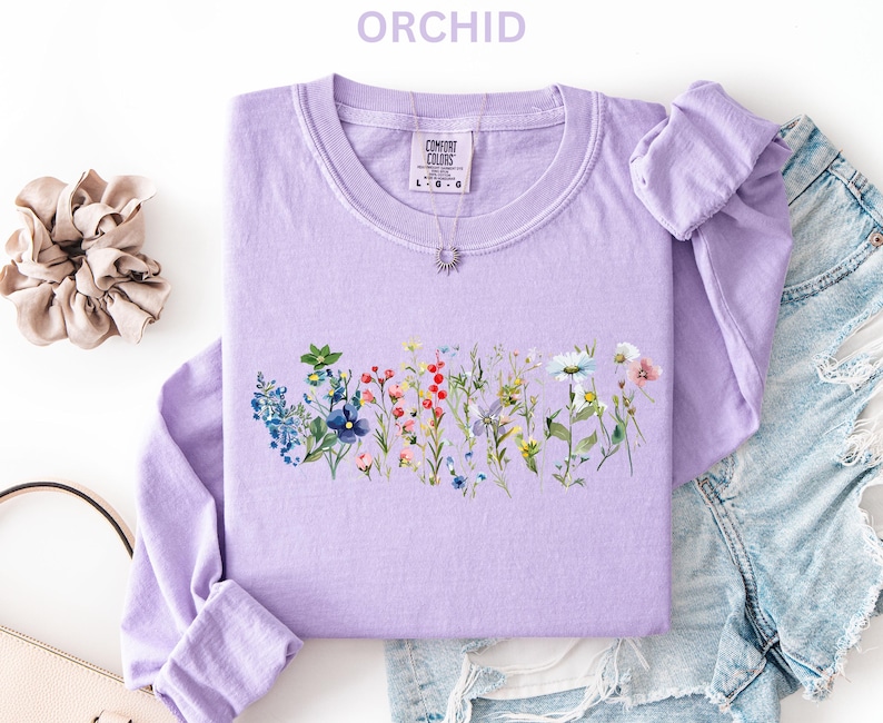 May include: A light purple long-sleeved sweatshirt with a floral design across the front. The design features a variety of colorful wildflowers. The word "ORCHID" is printed at the top. A scrunchie and a purse are also in the image.