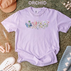 May include: A light purple t-shirt with the word "ORCHID" above the collar. The shirt features a floral design across the chest. The shirt is surrounded by a brown hat, white sneakers, and a blue blanket.
