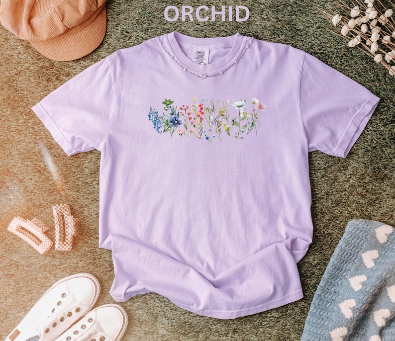 May include: A light purple t-shirt with the word "ORCHID" above the collar. The shirt features a floral design across the chest. The shirt is surrounded by a brown hat, white sneakers, and a blue blanket.