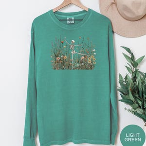 Puede incluir: Una camiseta de manga larga con cuello redondo en color verde azulado lavado. El centro de la prenda presenta una ilustraci&oacute;n gr&aacute;fica de un esqueleto humano de pie en un campo de flores silvestres coloridas, incluyendo margaritas y amapolas.