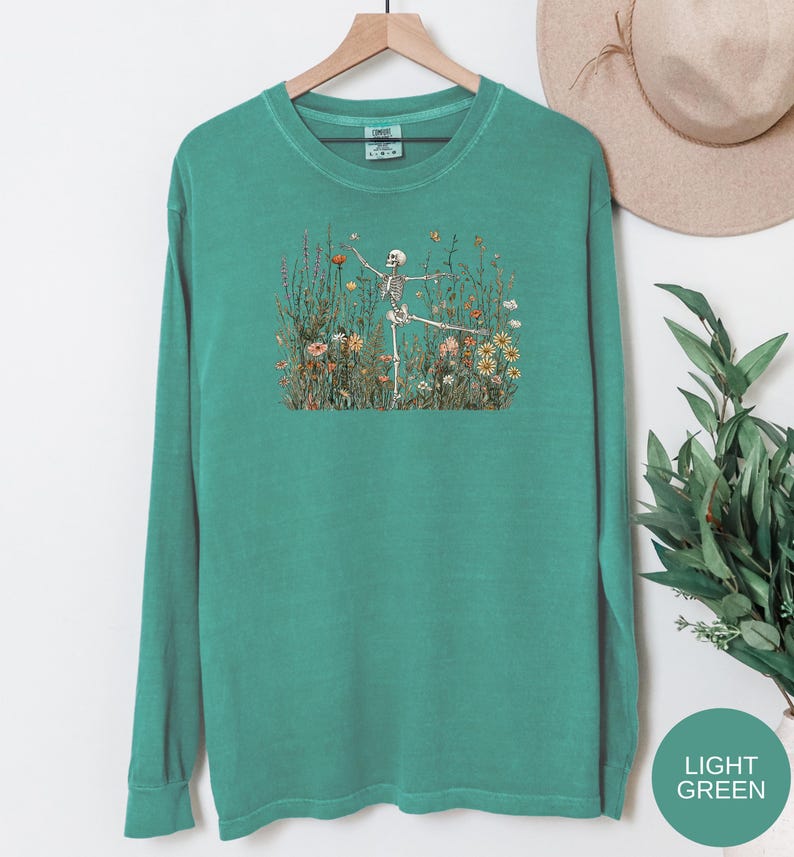 Puede incluir: Una camiseta de manga larga con cuello redondo en color verde azulado lavado. El centro de la prenda presenta una ilustraci&oacute;n gr&aacute;fica de un esqueleto humano de pie en un campo de flores silvestres coloridas, incluyendo margaritas y amapolas.