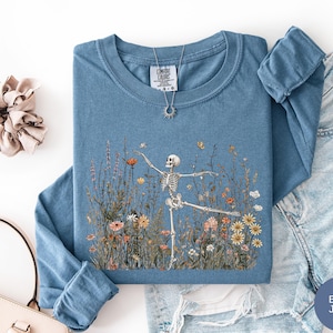Puede incluir: Una camiseta azul de manga larga con un gr&aacute;fico de un esqueleto bailando en un campo de flores silvestres coloridas. La camiseta tiene cuello redondo y una etiqueta Comfort Colors en el interior. Un collar con colgante de sol descansa sobre ella.