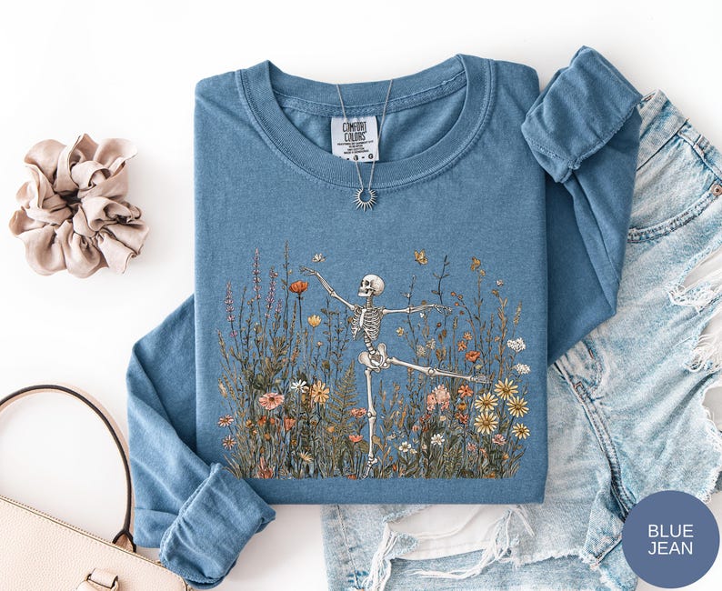 Puede incluir: Una camiseta azul de manga larga con un gr&aacute;fico de un esqueleto bailando en un campo de flores silvestres coloridas. La camiseta tiene cuello redondo y una etiqueta Comfort Colors en el interior. Un collar con colgante de sol descansa sobre ella.