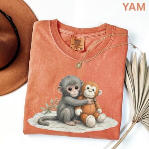 Puede incluir: Una camiseta Comfort Colors de color coral con un gr&aacute;fico de dos monos abraz&aacute;ndose. Uno es gris y el otro es un peluche. Un collar de oro con un colgante est&aacute; sobre la camiseta. La camiseta tiene cuello redondo y mangas cortas.