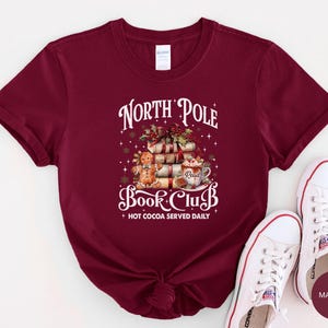 May include: A maroon short-sleeve t-shirt featuring a graphic of a gingerbread person, a stack of books, and a mug of hot cocoa. The text reads North Pole Book Club and Hot Cocoa Served Daily, surrounded by small white stars and snowflakes.