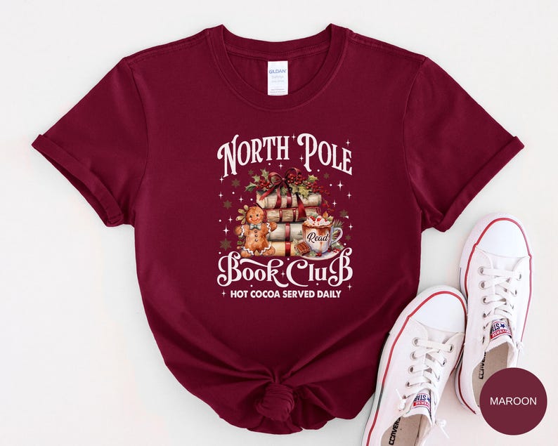 May include: A maroon short-sleeve t-shirt featuring a graphic of a gingerbread person, a stack of books, and a mug of hot cocoa. The text reads North Pole Book Club and Hot Cocoa Served Daily, surrounded by small white stars and snowflakes.