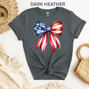May include: Dark heather gray t-shirt featuring a watercolor bow design in red, white, and blue, with stars and stripes. The bow is centered on the front of the shirt. The shirt is knotted at the waist.