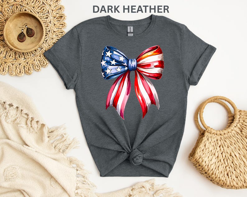 May include: Dark heather gray t-shirt featuring a watercolor bow design in red, white, and blue, with stars and stripes. The bow is centered on the front of the shirt. The shirt is knotted at the waist.