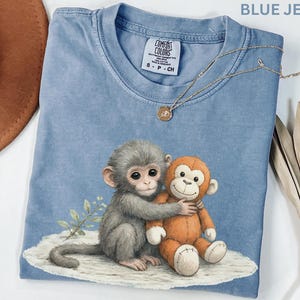 Puede incluir: Camiseta azul vaquero con un gr&aacute;fico de dos monos abraz&aacute;ndose. Un mono es gris, el otro es naranja. Un collar dorado con un peque&ntilde;o colgante descansa sobre la camisa. La camiseta tiene una etiqueta Comfort Colors.