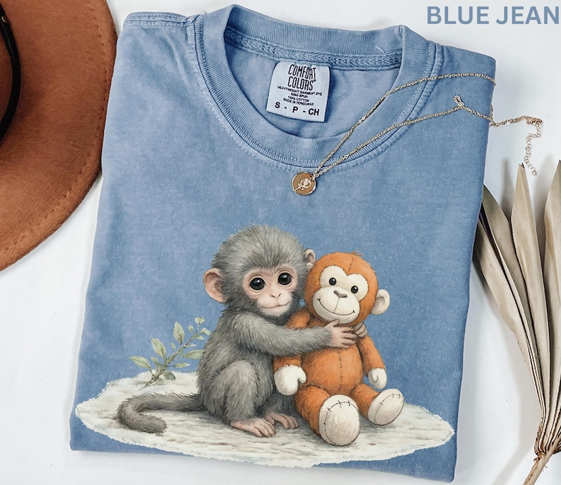 Puede incluir: Camiseta azul vaquero con un gr&aacute;fico de dos monos abraz&aacute;ndose. Un mono es gris, el otro es naranja. Un collar dorado con un peque&ntilde;o colgante descansa sobre la camisa. La camiseta tiene una etiqueta Comfort Colors.