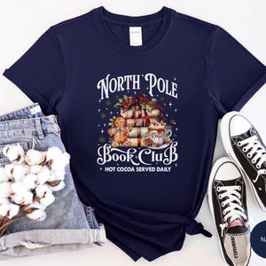 May include: A navy blue short-sleeve t-shirt featuring a graphic of a stack of books, a gingerbread person, and a mug of hot cocoa. The text reads North Pole Book Club and Hot Cocoa Served Daily. The design includes holly, stars, and snowflakes.