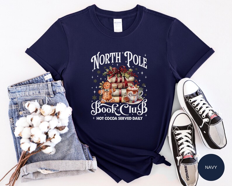 May include: A navy blue short-sleeve t-shirt featuring a graphic of a stack of books, a gingerbread person, and a mug of hot cocoa. The text reads North Pole Book Club and Hot Cocoa Served Daily. The design includes holly, stars, and snowflakes.