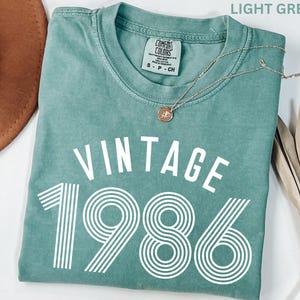 May include: Light green t-shirt with the text "VINTAGE 1986" in white. The shirt has a crew neck and short sleeves. A silver necklace with a pendant is draped across the shirt.