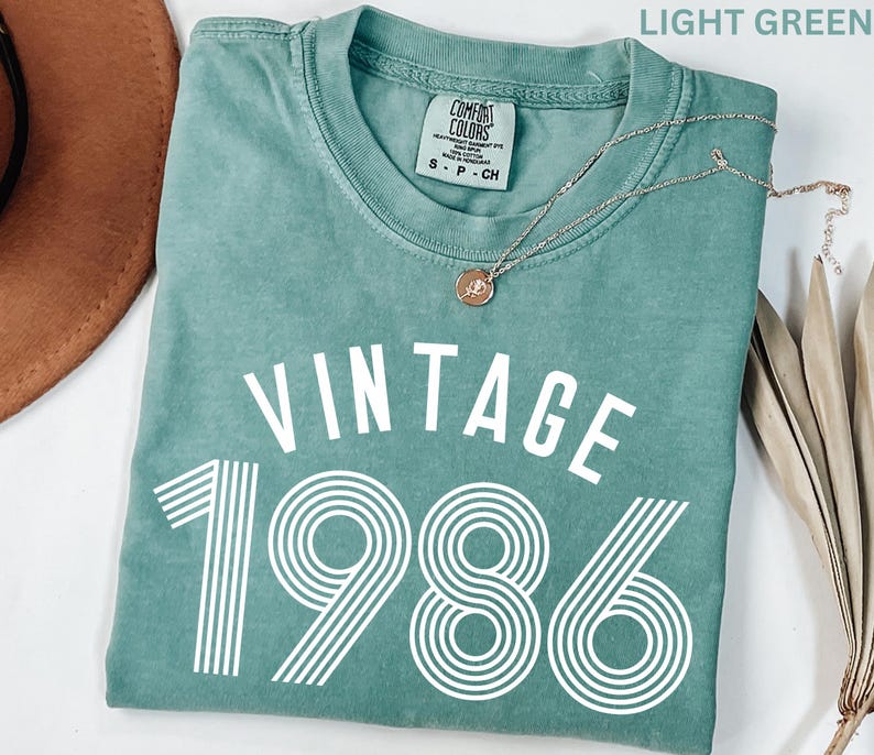 May include: Light green t-shirt with the text "VINTAGE 1986" in white. The shirt has a crew neck and short sleeves. A silver necklace with a pendant is draped across the shirt.