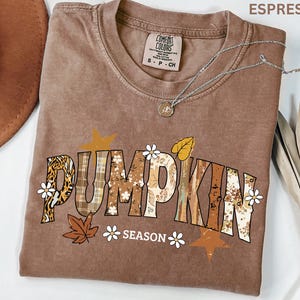 May include: An espresso-colored short-sleeve t-shirt with a graphic print that reads PUMPKIN SEASON. The letters feature various patterns including leopard print, plaid, and floral designs, accented with small white flowers and brown leaves.