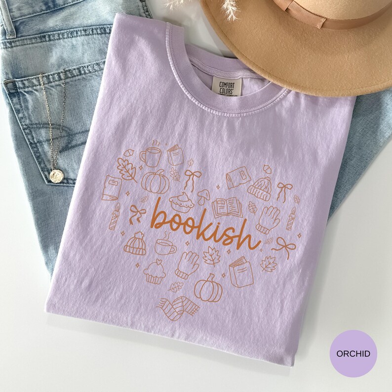 May include: A light purple t-shirt with a crew neckline and a Comfort Colors label. The front features a heart-shaped graphic composed of brown line-art icons including books, pumpkins, mugs, mittens, and bows, with the word bookish in the center.