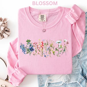 May include: A light pink long-sleeved shirt with a floral design across the chest. The shirt has a crew neck and the word "BLOSSOM" is printed at the top. A scrunchie, purse, and denim shorts are also in the image.