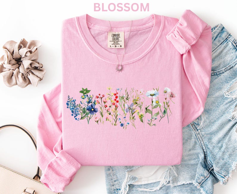 May include: A light pink long-sleeved shirt with a floral design across the chest. The shirt has a crew neck and the word "BLOSSOM" is printed at the top. A scrunchie, purse, and denim shorts are also in the image.