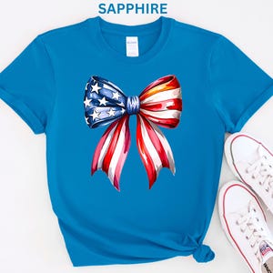 May include: Sapphire blue t-shirt featuring a watercolor bow design in red, white, and blue, with stars. The bow is the focal point, evoking patriotic themes. White canvas sneakers with red accents are placed near the shirt.