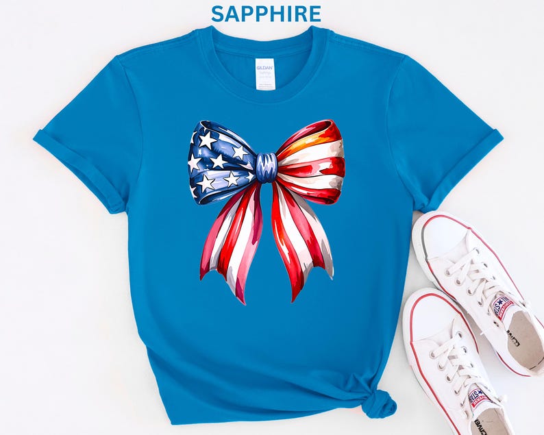 May include: Sapphire blue t-shirt featuring a watercolor bow design in red, white, and blue, with stars. The bow is the focal point, evoking patriotic themes. White canvas sneakers with red accents are placed near the shirt.