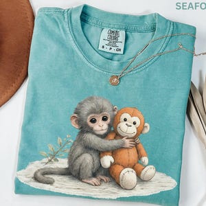 Puede incluir: Camiseta Comfort Colors verde mar con un dibujo de un mono gris abrazando a un mono de peluche naranja. La camiseta tiene cuello redondo y mangas cortas. Un collar dorado con un peque&ntilde;o colgante est&aacute; drapeado sobre la camiseta.