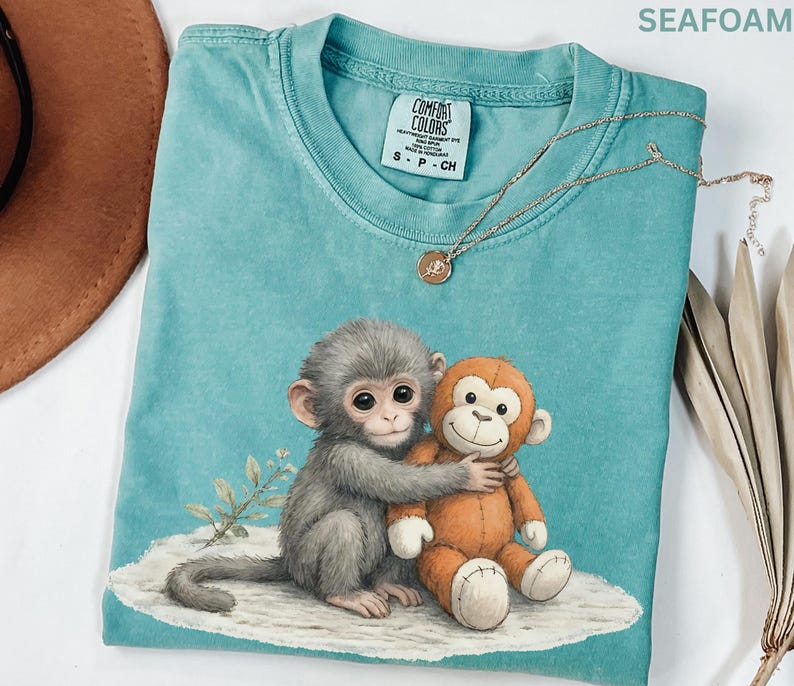 Puede incluir: Camiseta Comfort Colors verde mar con un dibujo de un mono gris abrazando a un mono de peluche naranja. La camiseta tiene cuello redondo y mangas cortas. Un collar dorado con un peque&ntilde;o colgante est&aacute; drapeado sobre la camiseta.