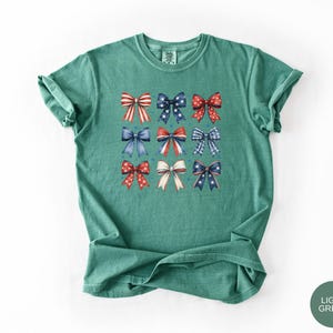 May include: A light green short-sleeve t-shirt featuring a grid of nine decorative bows in various red, white, and blue patterns, including stripes, polka dots, and stars, arranged in three rows on the center of the chest.
