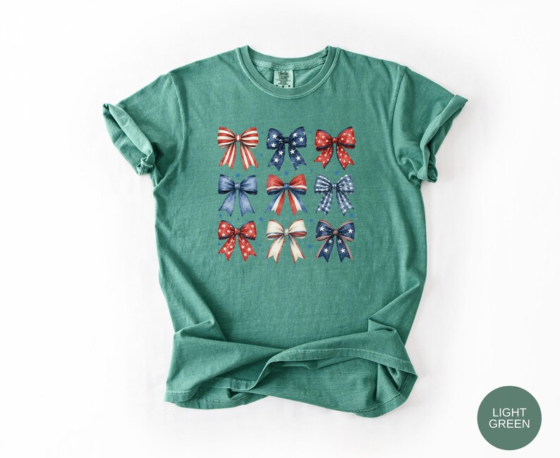 May include: A light green short-sleeve t-shirt featuring a grid of nine decorative bows in various red, white, and blue patterns, including stripes, polka dots, and stars, arranged in three rows on the center of the chest.