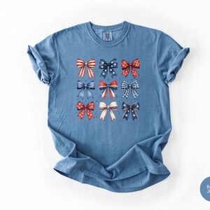 May include: A blue jean colored short-sleeve t-shirt featuring a graphic print of nine decorative bows arranged in a grid. The bows display various patterns including red and white stripes, blue with white stars, and red with white polka dots.