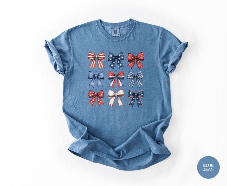 May include: A blue jean colored short-sleeve t-shirt featuring a graphic print of nine decorative bows arranged in a grid. The bows display various patterns including red and white stripes, blue with white stars, and red with white polka dots.