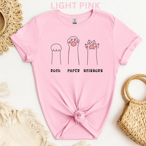 Puede incluir: Camiseta rosa claro con un dise&ntilde;o l&uacute;dico de patas de gato que representan piedra, papel y tijeras. El texto "ROCK PAPER SCISSORS" est&aacute; impreso en negro debajo. La camiseta est&aacute; hecha de material suave.