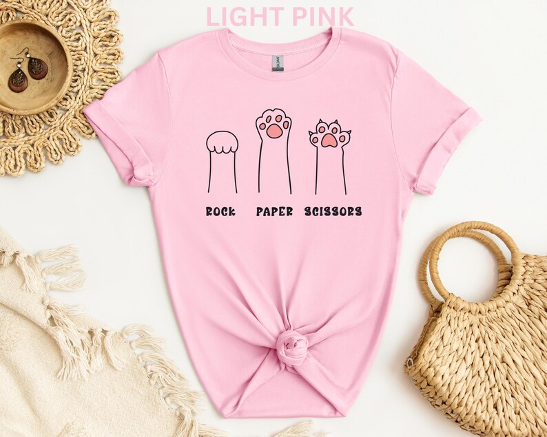 Puede incluir: Camiseta rosa claro con un dise&ntilde;o l&uacute;dico de patas de gato que representan piedra, papel y tijeras. El texto "ROCK PAPER SCISSORS" est&aacute; impreso en negro debajo. La camiseta est&aacute; hecha de material suave.
