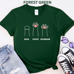 Puede incluir: Camiseta verde bosque con un dise&ntilde;o l&uacute;dico de "Piedra, Papel, Tijeras" con patas de gato. El dise&ntilde;o incluye tres ilustraciones de patas que representan cada elemento del juego. La camiseta se combina con vaqueros azules y zapatillas negras.