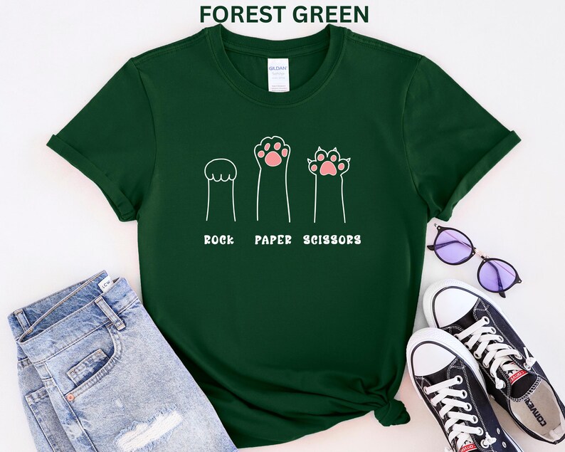 Puede incluir: Camiseta verde bosque con un dise&ntilde;o l&uacute;dico de "Piedra, Papel, Tijeras" con patas de gato. El dise&ntilde;o incluye tres ilustraciones de patas que representan cada elemento del juego. La camiseta se combina con vaqueros azules y zapatillas negras.