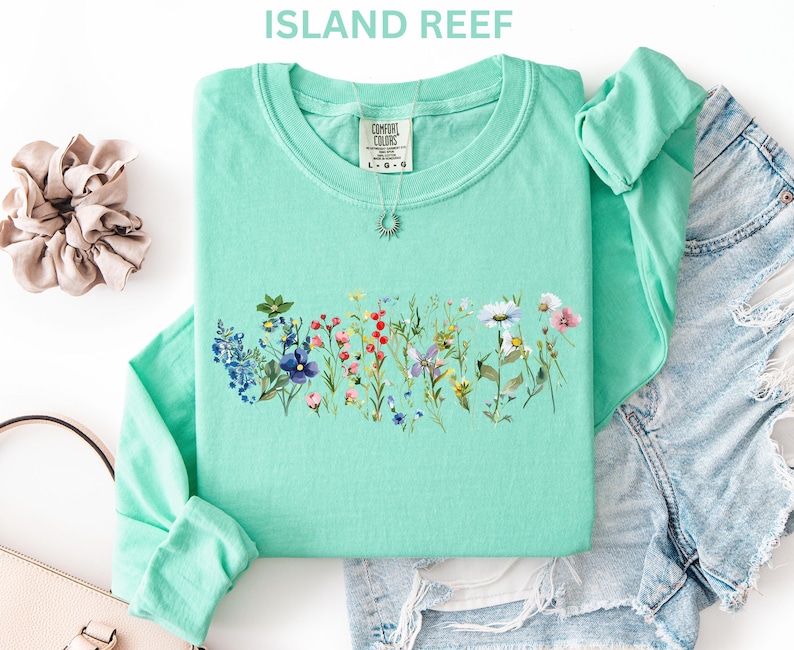 May include: A mint green long-sleeved shirt with a floral design across the chest. The design features a variety of colorful wildflowers. The shirt is folded with a pair of denim shorts and a scrunchie. The text "ISLAND REEF" is at the top.