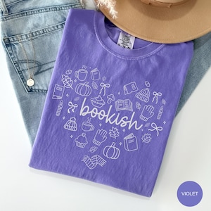 May include: A violet short-sleeved t-shirt featuring a white line-art graphic of various autumn and reading-themed icons arranged in a heart shape, with the word "bookish" written in cursive script across the center of the design.