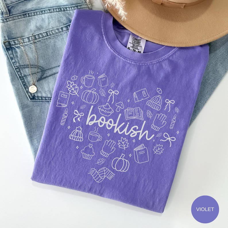 May include: A violet short-sleeved t-shirt featuring a white line-art graphic of various autumn and reading-themed icons arranged in a heart shape, with the word "bookish" written in cursive script across the center of the design.