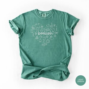 May include: A light green short-sleeve t-shirt featuring a white line-art graphic in the shape of a heart. The design includes small illustrations of books, pumpkins, mugs, and leaves, with the word bookish written in cursive in the center.