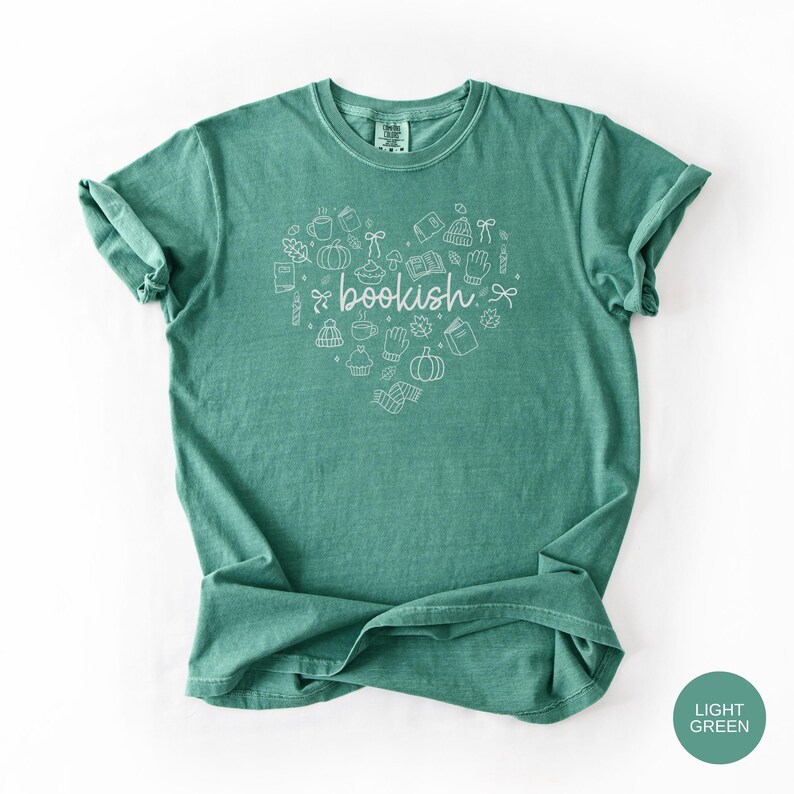 May include: A light green short-sleeve t-shirt featuring a white line-art graphic in the shape of a heart. The design includes small illustrations of books, pumpkins, mugs, and leaves, with the word bookish written in cursive in the center.