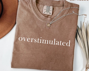 Puede incluir: Una camiseta de manga corta de color marrón con la palabra overstimulated impresa en blanco en el pecho. La camiseta cuenta con una etiqueta de la marca Comfort Colors en el cuello interior y se muestra con un collar plateado y un sombrero marrón.