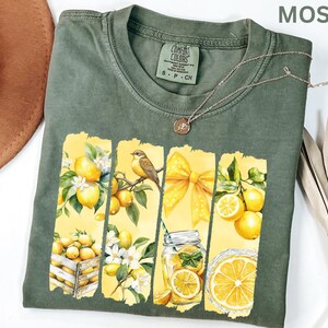 May include: Moss green t-shirt with a lemon-themed graphic. The design features lemons, a bird, a yellow bow, and a jar of lemonade. A silver necklace with a pendant is on the shirt. The shirt is labeled "Comfort Colors".