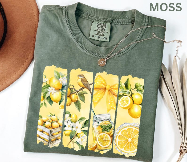May include: Moss green t-shirt with a lemon-themed graphic. The design features lemons, a bird, a yellow bow, and a jar of lemonade. A silver necklace with a pendant is on the shirt. The shirt is labeled "Comfort Colors".