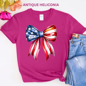 May include: A magenta t-shirt featuring a watercolor bow design with the American flag's stars and stripes. The bow is centered on the shirt, with the text "ANTIQUE HELICONIA" at the top. Blue jeans are visible on the right.