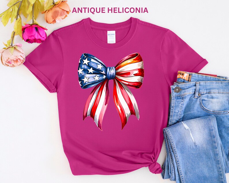 May include: A magenta t-shirt featuring a watercolor bow design with the American flag's stars and stripes. The bow is centered on the shirt, with the text "ANTIQUE HELICONIA" at the top. Blue jeans are visible on the right.